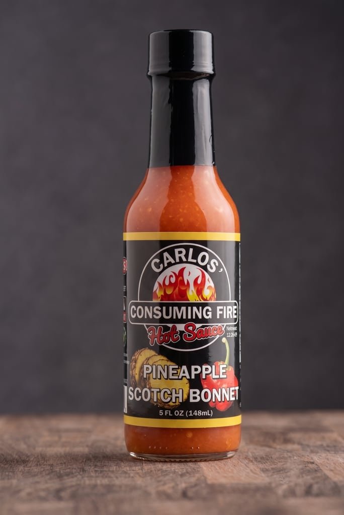 Carlos' Consuming Fire Hot Sauce Pineapple Scotch Bonnet bottle, 5 FL OZ, on a wooden surface.