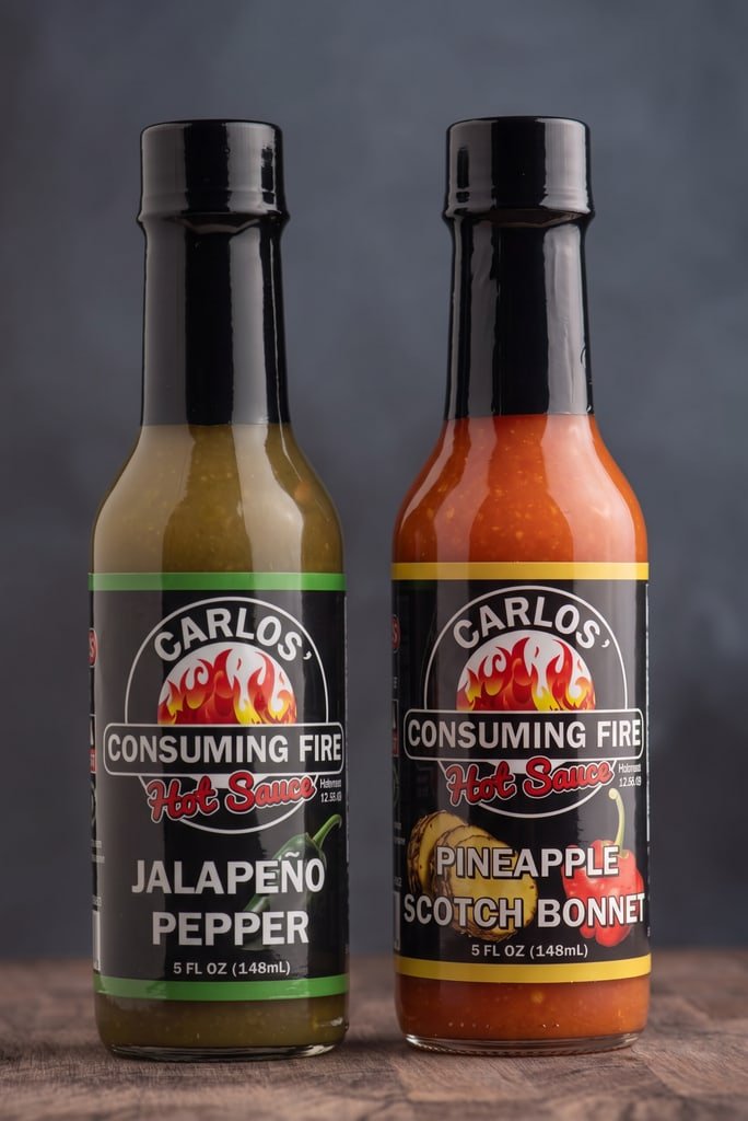 Carlos' Consuming Fire Hot Sauce Twice as Nice Bundle: Jalapeño Pepper and Pineapple Scotch Bonnet hot sauce bottles on a rustic wooden surface.