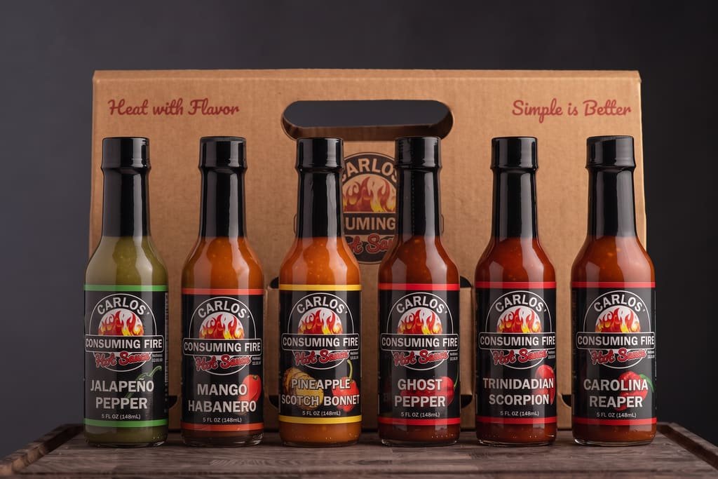 Carlos' Consuming Fire Hot Sauce Ultimate Bundle: six hot sauce bottles — Jalapeño Pepper, Mango Habanero, Pineapple Scotch Bonnet, Ghost Pepper, Trinidadian Scorpion and Carolina Reaper — in front of a kraft gift carrier.