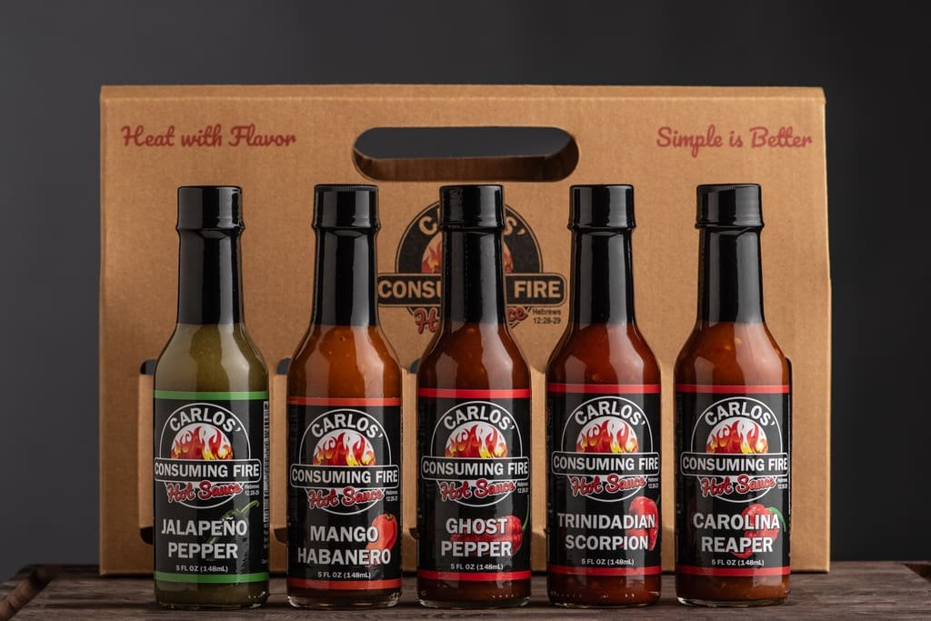 A professional lineup of five Carlos' Consuming Fire Hot Sauce bottles ranging from Jalapeno to Carolina Reaper, set against a gift carrier box.